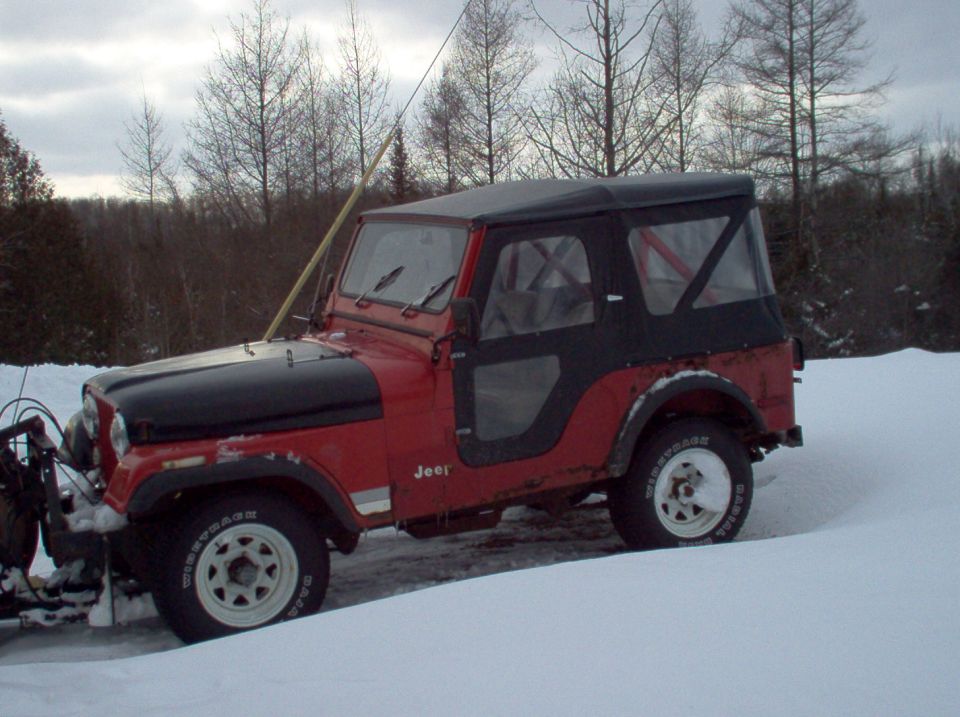 Red Jeep CJ-5 with black soft top and snow plow parked on snow under a gray winter sky