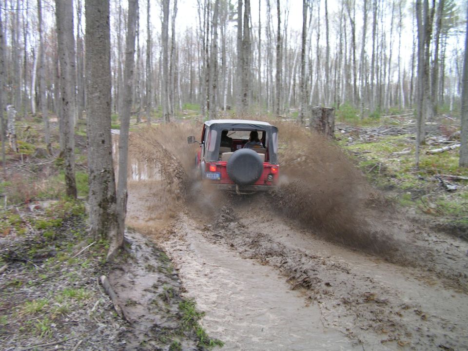 red Jeep Wrangler driving through mud and splashing water on a wooded trail
