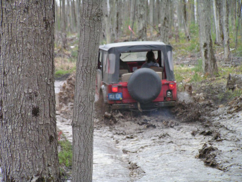Red Jeep CJ-series vehicle with a spare tire on the back, driving through a muddy forest path