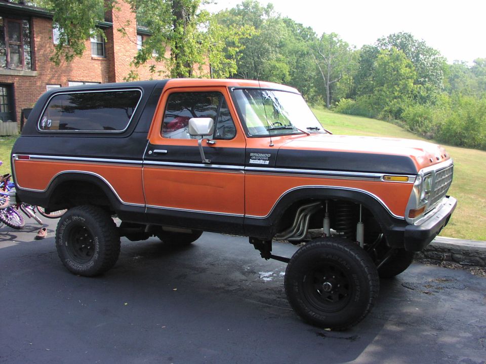 Orange and black lifted Ford Bronco with custom exhaust pipes visible under the front fender. Tires are large and black