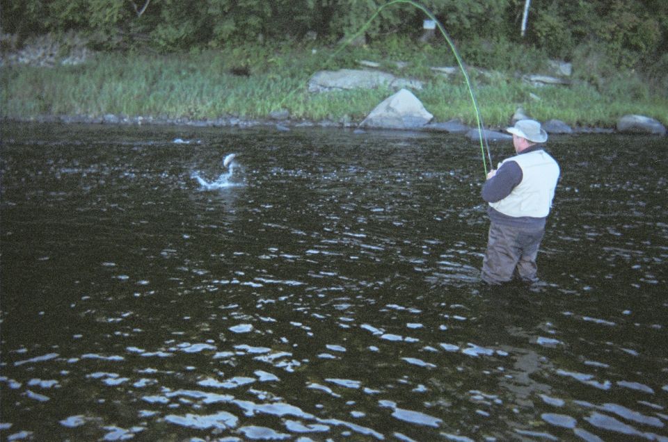 Man in waders casts fishing line into choppy river water with trees and rocks on the bank