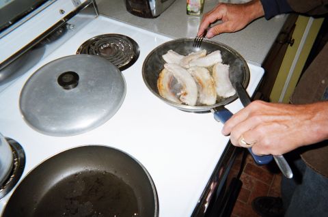 1984 Ford Bronco - Someone is cooking bacon on a stovetop, with a fork and tongs in hand.