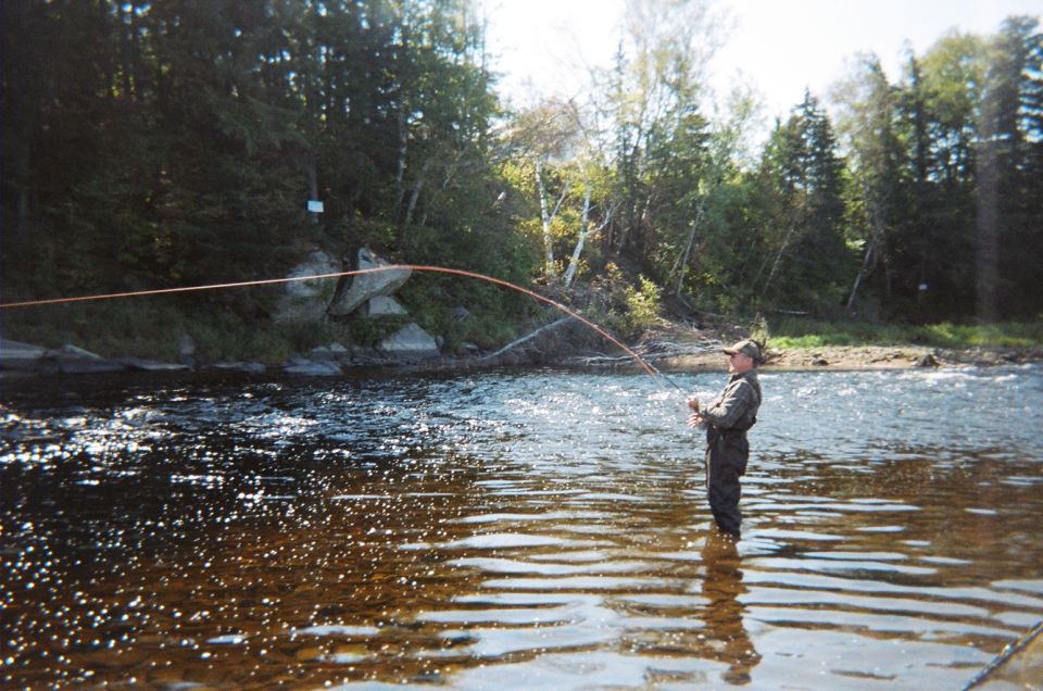 Man wearing waders and a hat, fly fishing in a river with trees and rocks in the background