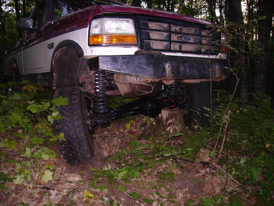 Maroon and white Ford F-Series truck with lifted suspension, front view, off-road tires, in wooded area