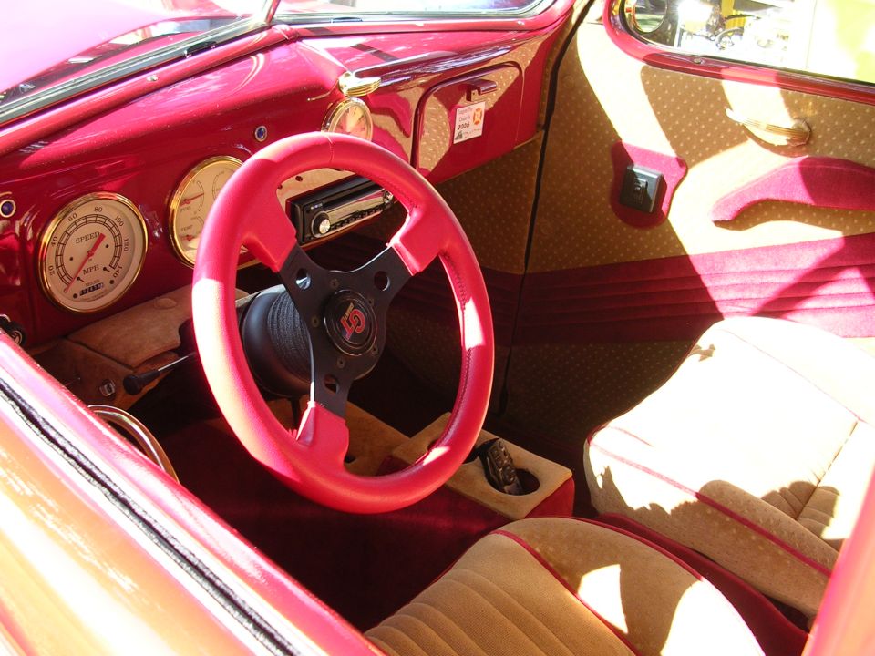 Red steering wheel and tan interior of a classic car. Visible gauges and radio