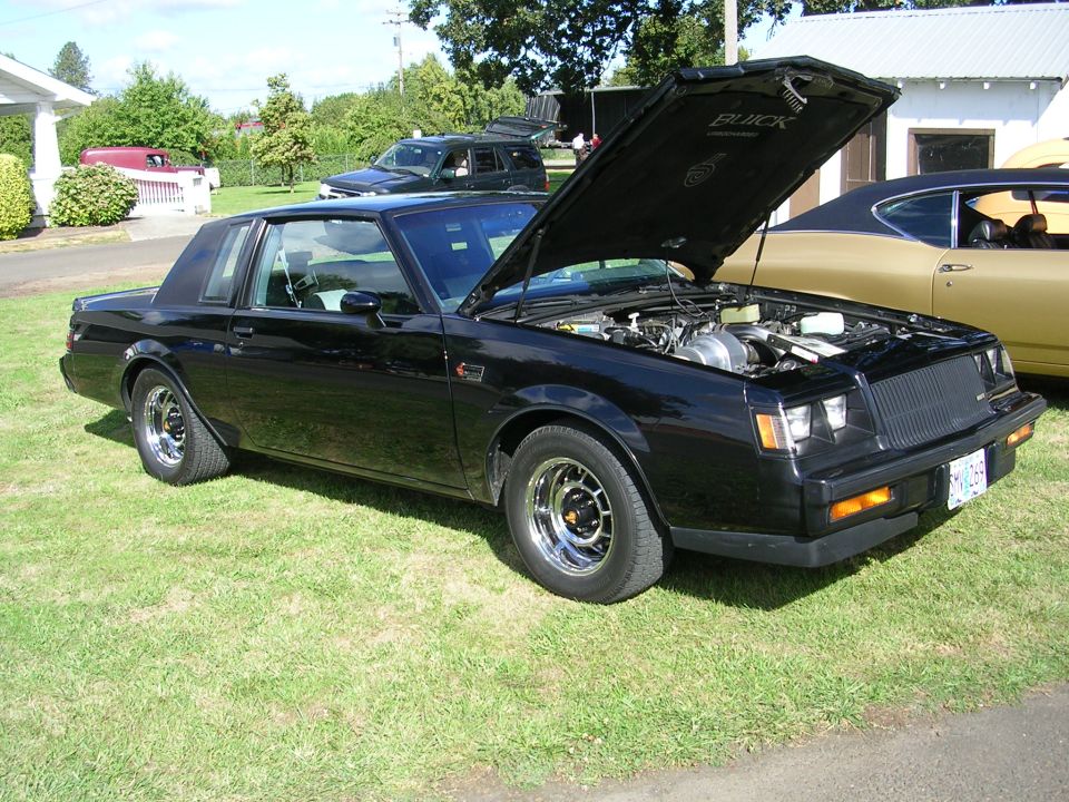 Black Buick Grand National with hood open, parked on grass. Another car is visible to the right