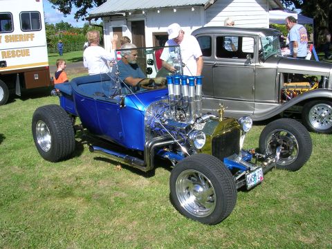 1996 Ford Probe - A custom blue hot rod with a large V8 engine is parked on grass next to a silver sedan.