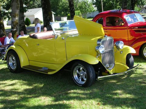 1996 Ford Probe - A bright yellow convertible hot rod is displayed with its hood open, parked next to a red car at an outdoor car show.