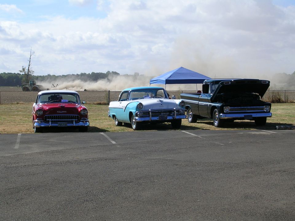Red car, blue and white car, and black pickup truck with hood open parked in lot. Tractor working in field behind them