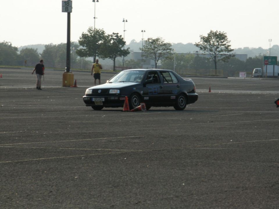 Dark sedan with racing cones on asphalt lot, two people in background
