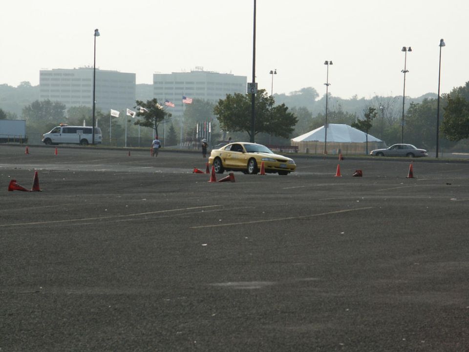 Yellow coupe with a rounded front end and a low profile, seen from a 3/4 front angle on a paved lot with orange cones