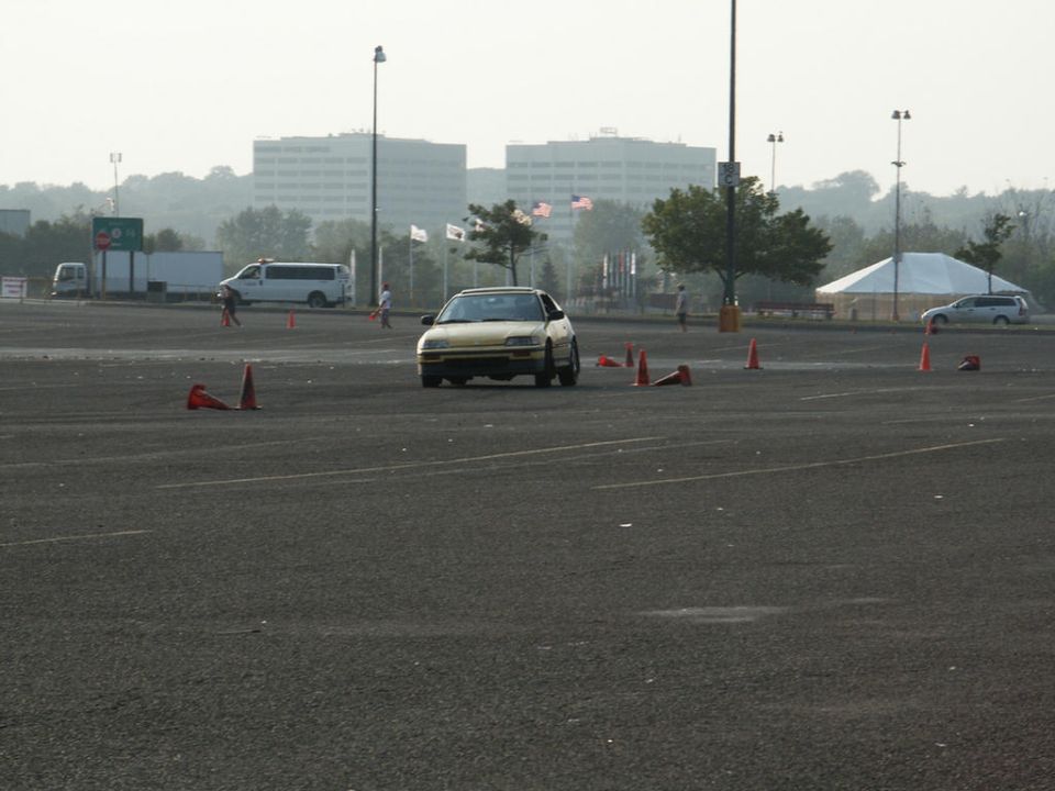 Yellow sedan on asphalt with orange cones, buildings and tents in background