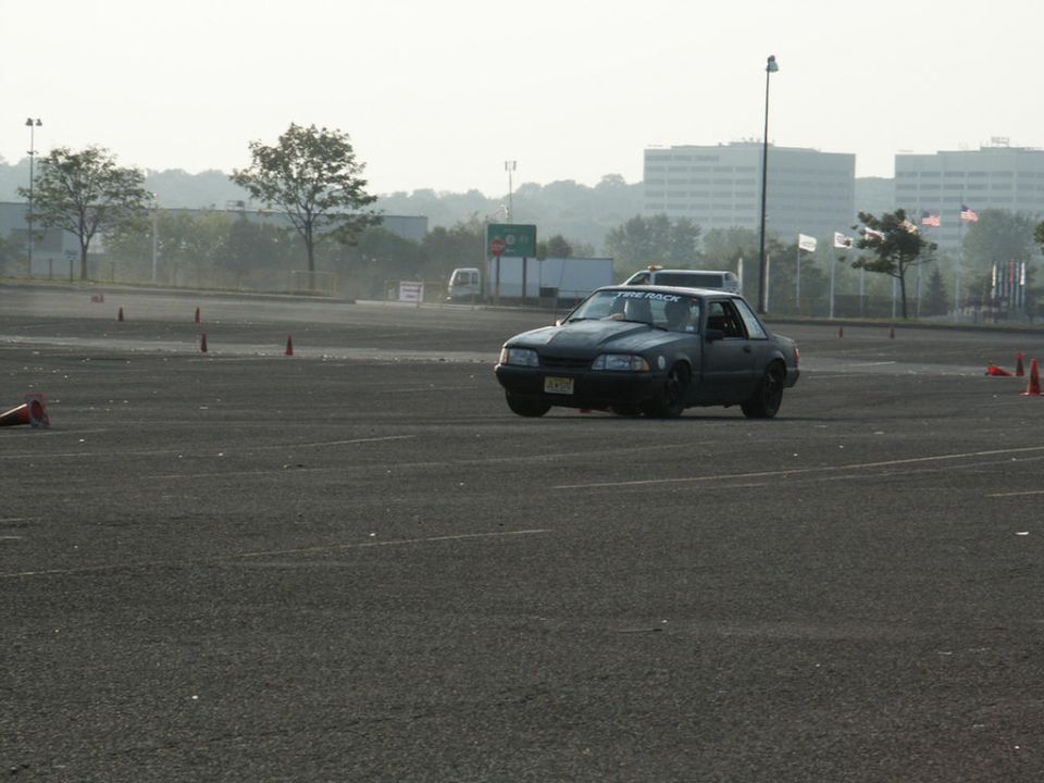 Dark coupe with aftermarket hood scoop and "TIRE RACK" sticker on windshield, driving on asphalt course with cones
