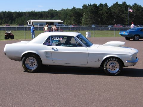 1989 Ford Mustang - A white classic Mustang with racing slicks and a hood scoop is parked at a drag strip, with other vehicles and spectators in the background.