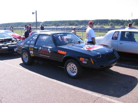 1989 Ford Mustang - A black coupe with racing decals and "Turbo" text is parked at a drag strip, with other vehicles visible in the background.