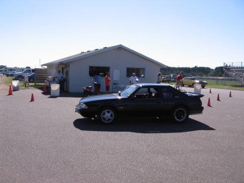1989 Ford Mustang - A black 1989 Ford Mustang is parked near a building at a race track, with other vehicles and people in the background.
