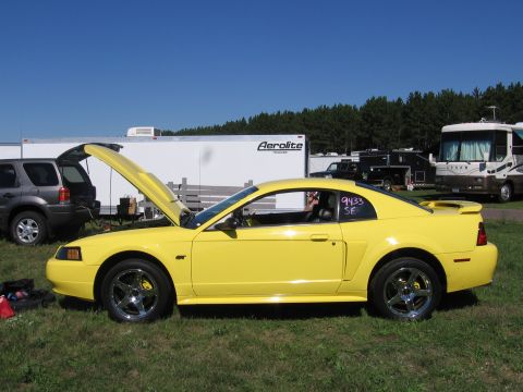 1989 Ford Mustang - A yellow coupe sits on grass with its hood open, parked near trailers and other vehicles under a clear blue sky.