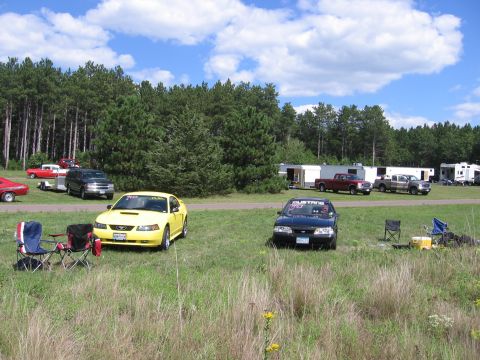 1989 Ford Mustang - A yellow Ford Mustang is parked on a grassy field with other vehicles and trailers in the background.