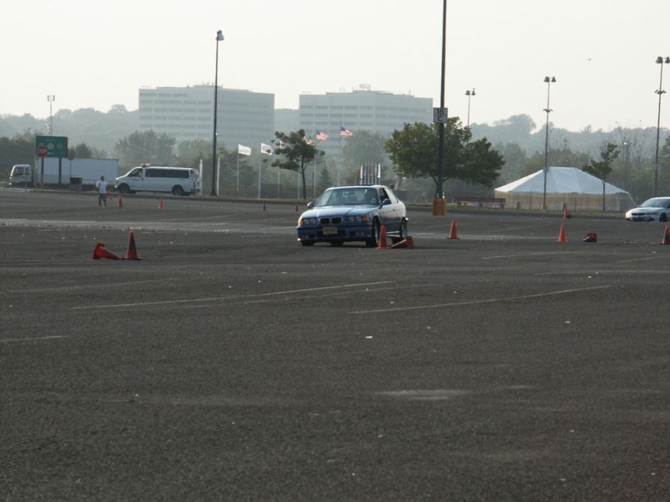 BMW E36 coupe navigating a course marked by orange cones, with vans and buildings in the background