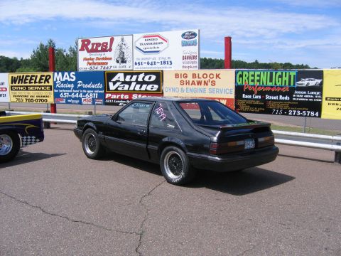 1989 Ford Mustang - A black Ford Mustang hatchback with racing numbers on the window is parked at a drag strip.