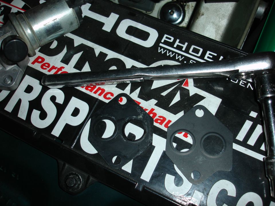Two exhaust manifold gaskets and a solenoid valve sit on a black mat with white text. A ratchet is also visible