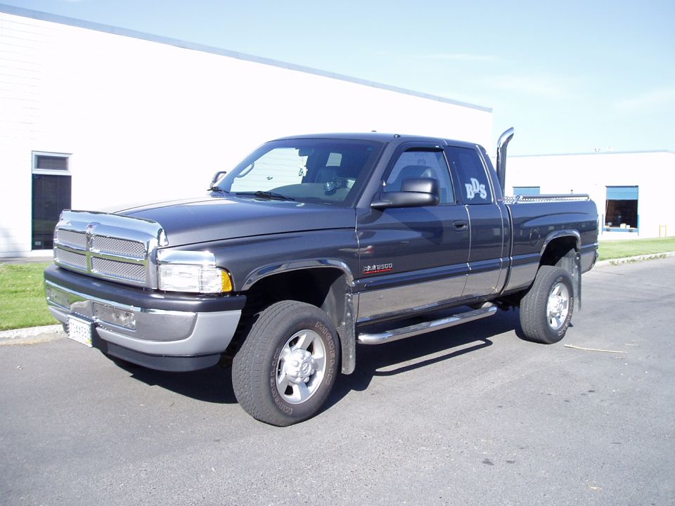 Dark gray Dodge Ram 2500 extended-cab pickup, front-left three-quarter view, parked outside industrial buildings.
