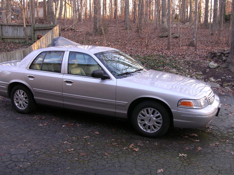 Silver Ford Crown Victoria sedan, passenger-side profile, parked on driveway near wooded backyard