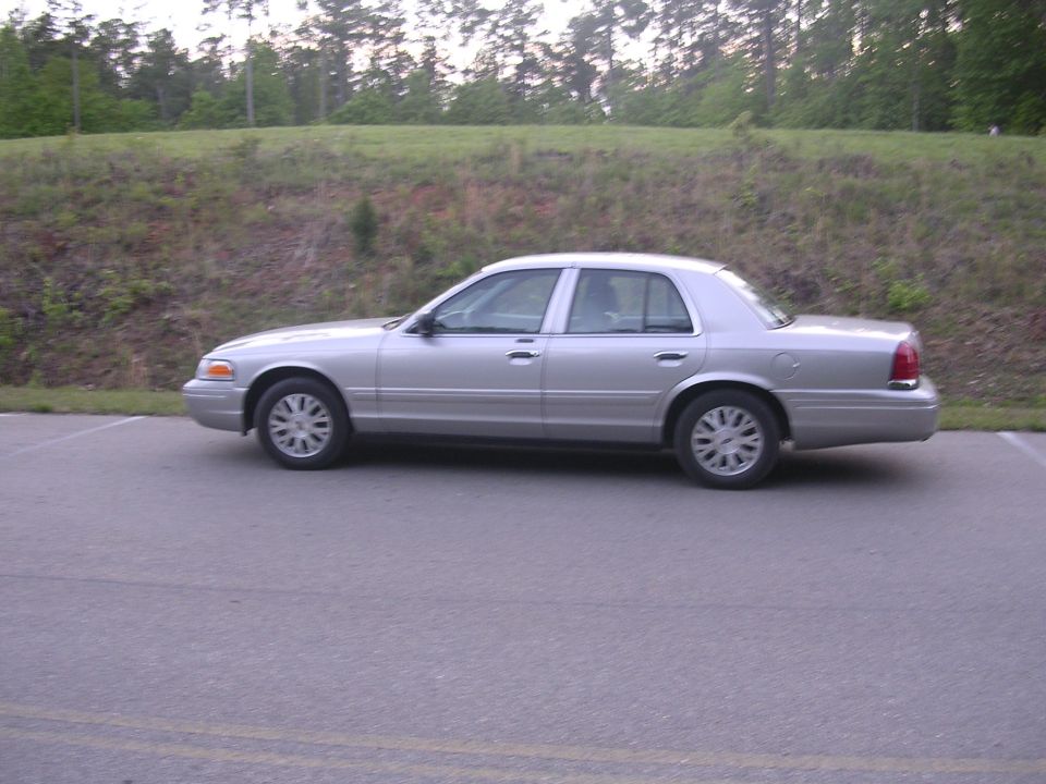 Silver Ford Crown Victoria sedan, driver-side profile, parked in lot beside grassy slope