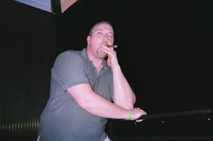 1984 Ford Bronco - A man enjoys a cigar while leaning on a railing against a dark background.