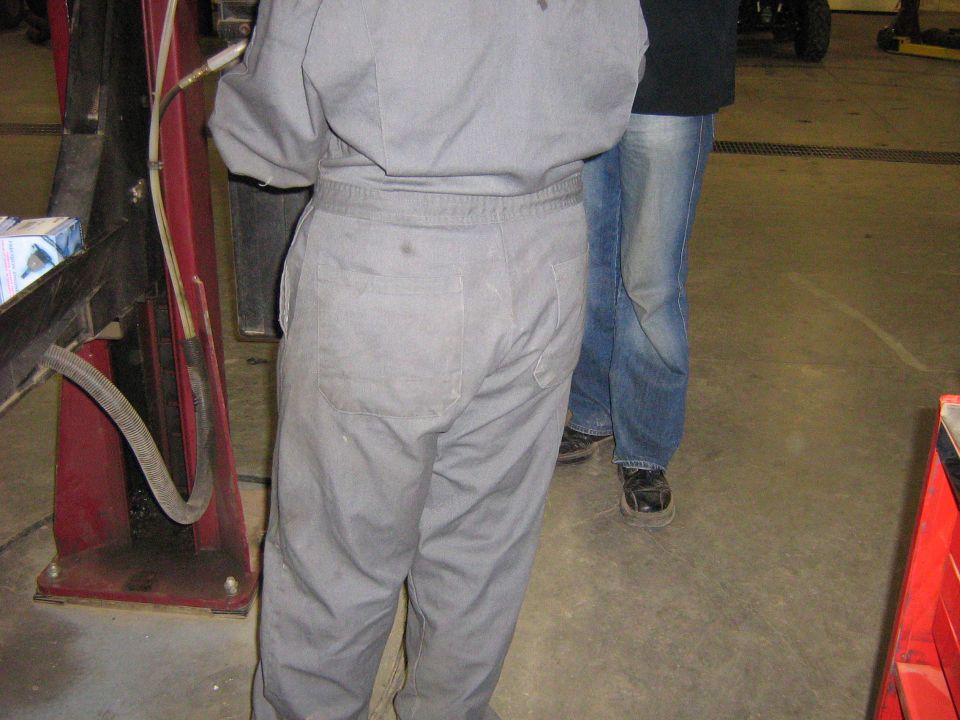 Two people standing in a garage, one in gray work clothes, the other in jeans. A red lift is visible