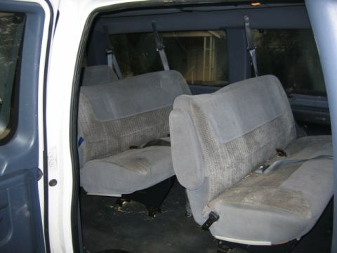1994 Ford Bronco - The interior of a van shows two rows of gray cloth seats with seatbelts. The floor is dark and appears dirty.