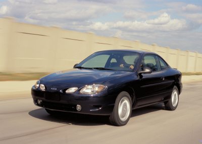2002 Ford Escort - A black 2002 Ford Escort ZX2 coupe is captured in motion on a road with a beige barrier in the background.