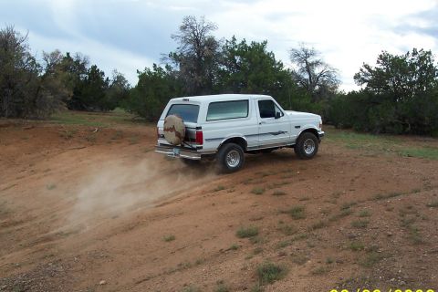 1996 Ford Bronco - A white Ford Bronco drives off-road, creating dust clouds as it navigates the terrain.