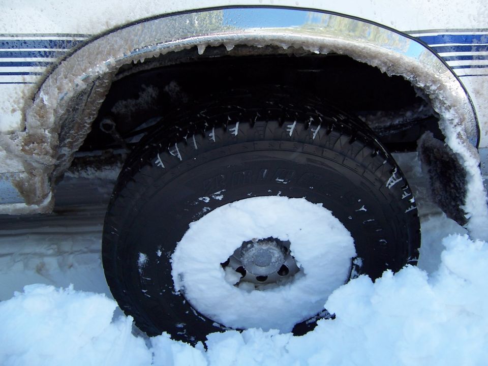 Close-up of a vehicle's front wheel well and tire, covered in ice and snow. Blue stripe visible on the body