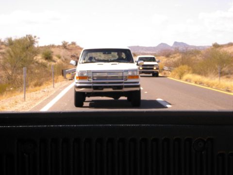 1996 Ford Bronco - Two trucks drive down a highway with desert scenery under a bright sky.
