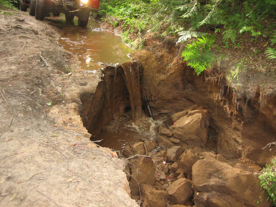 Front tires of an off-road vehicle at the edge of a muddy, eroded trail with water cascading into a rocky gully.