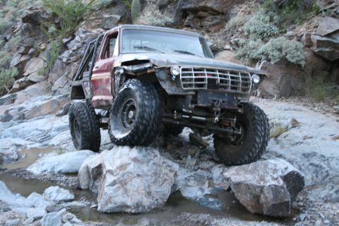 1966 Ford Bronco - Modified off-road vehicle navigates a rocky, dry riverbed with significant body damage and a custom roll cage.