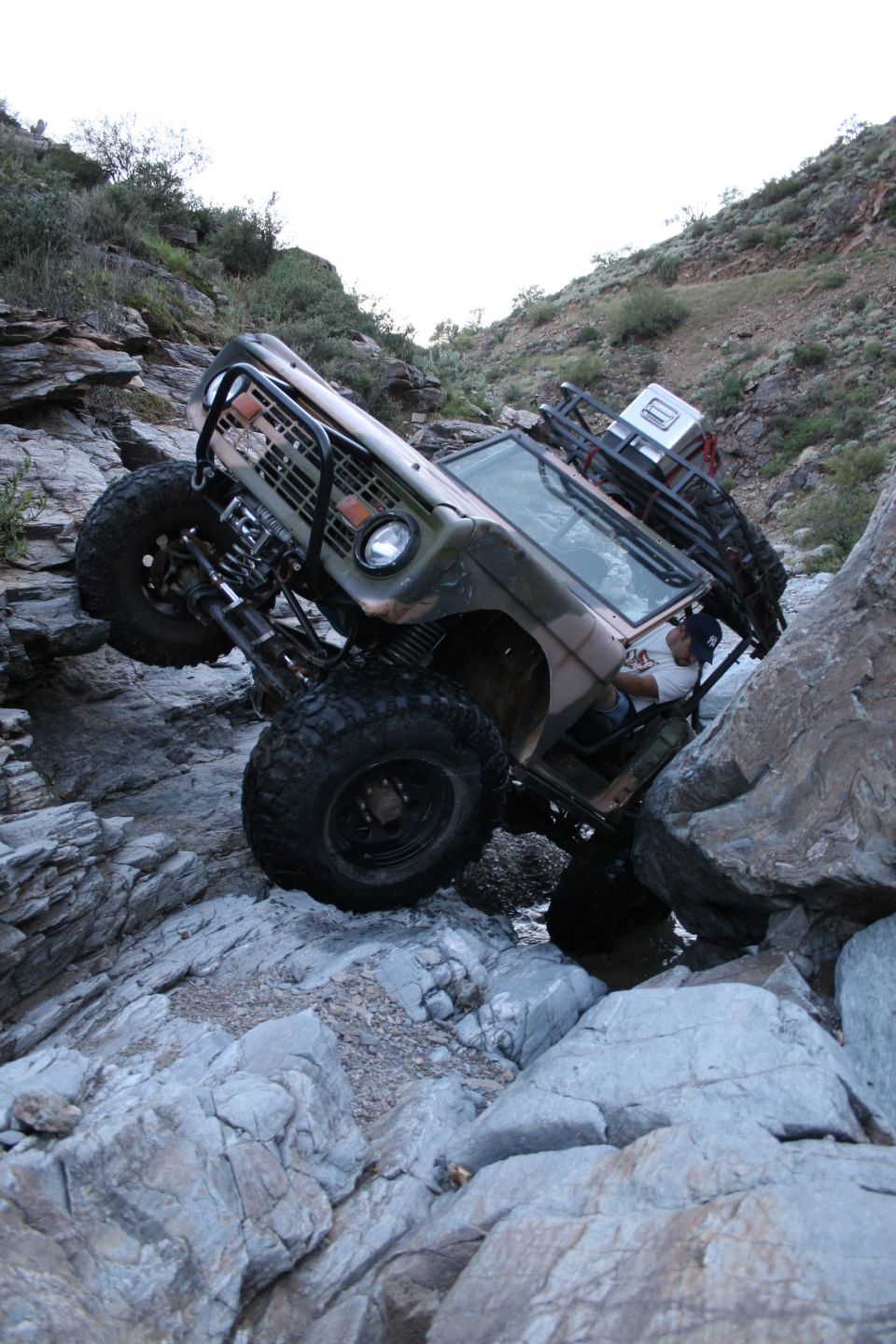 Camo doorless Bronco flexed sideways on a rock ledge, winch bumper and 35s visible