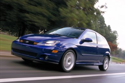 2002 Ford Focus - A blue two-door hatchback car is captured in motion on a road, with trees blurred in the background.