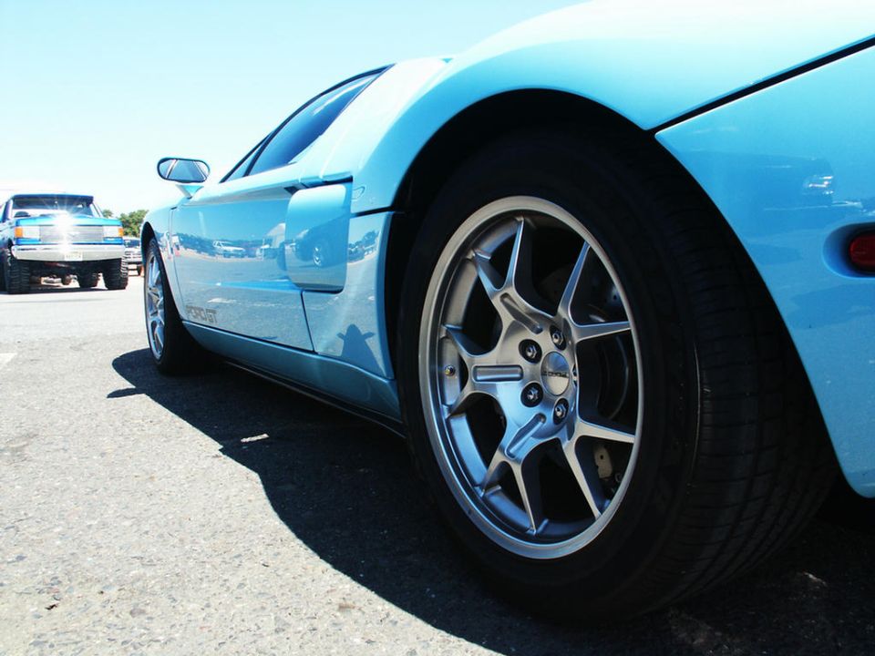 Close-up of a blue sports car's wheel and side panel, with a pickup truck in the background