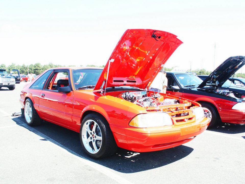 Red coupe with hood open, showing engine. White five-spoke wheels. Another car with hood open behind it