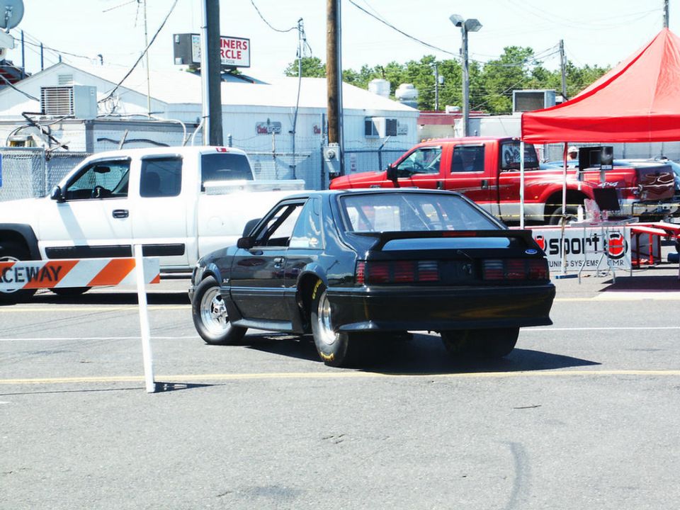 Black coupe with spoiler, rear view. White and red trucks in background