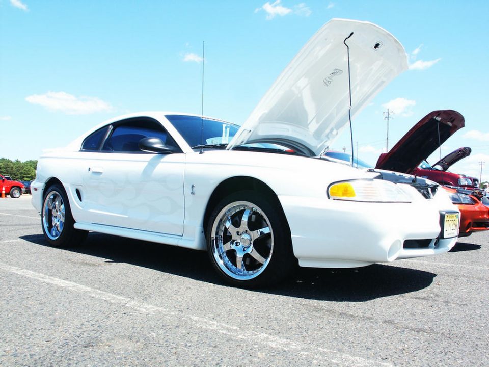 White Ford Mustang with flames on the side, hood up. Other cars with hoods up in background