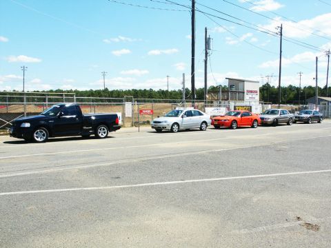 2001 Mercury Grand Marquis - A line of vehicles, including a black pickup truck and several cars, are parked near a race track on a sunny day.