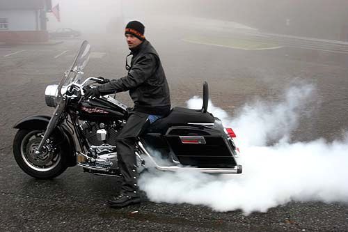 Man on black Harley-Davidson motorcycle doing a burnout in a parking lot, creating a large cloud of smoke