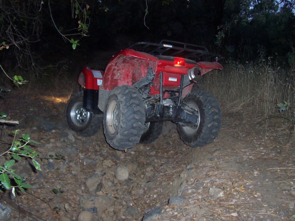 Red ATV on rocky terrain, rear view, with large knobby tires and a cargo rack
