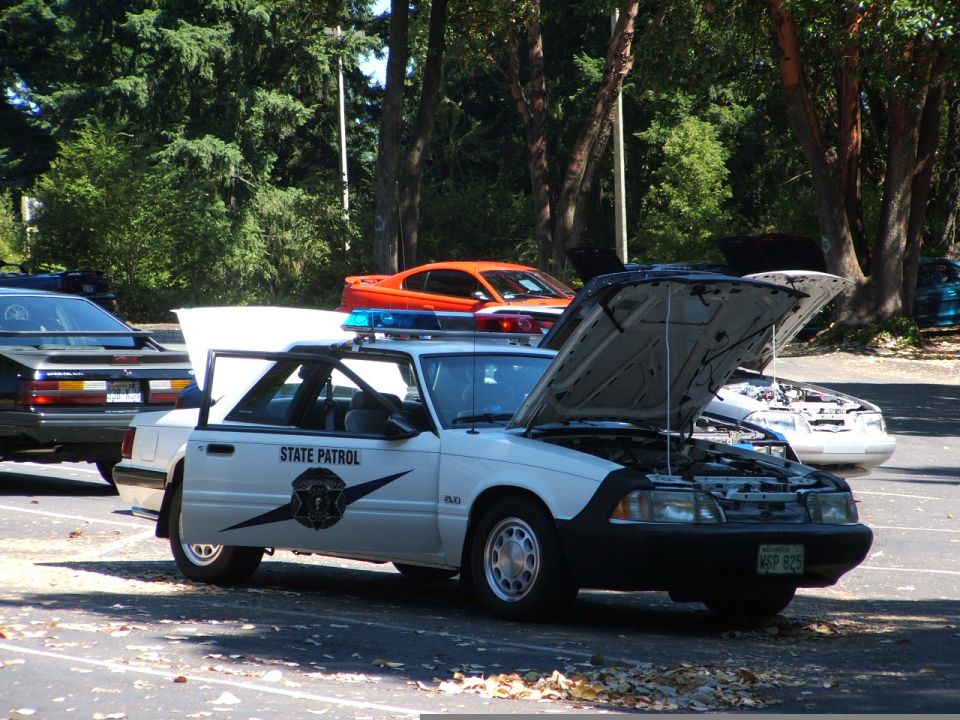 White 1987 Ford Mustang police car, side view, with "STATE PATROL" text and blue lights