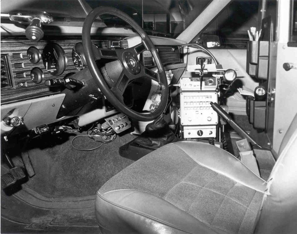 B&W interior of a police car showing steering wheel, woodgrain dash, and center-mounted radio console with mic.