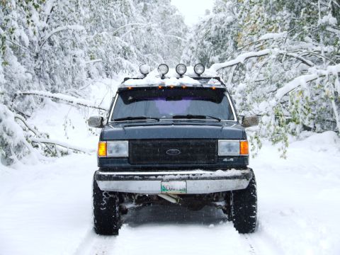 1988 Ford Bronco - A dark blue Ford Bronco navigates a snow-covered forest road, equipped with four auxiliary lights on its roof.