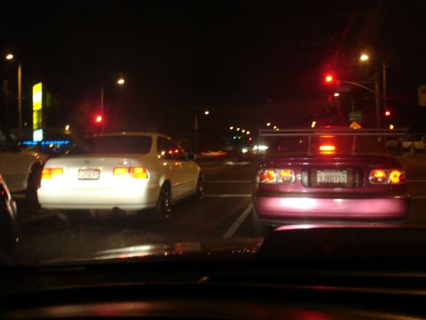 1993 Ford Taurus - Two cars are stopped at a traffic light at night, one purple with a spoiler and one white.